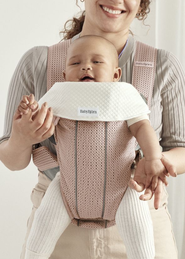 A smiling adult holds a baby in a pink BabyBjörn carrier. The baby wears a white bib and is dressed in a pink outfit with white leggings.