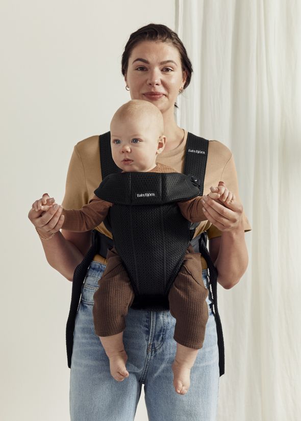 Woman holding a baby in a black BabyBjörn carrier, both wearing neutral tones.