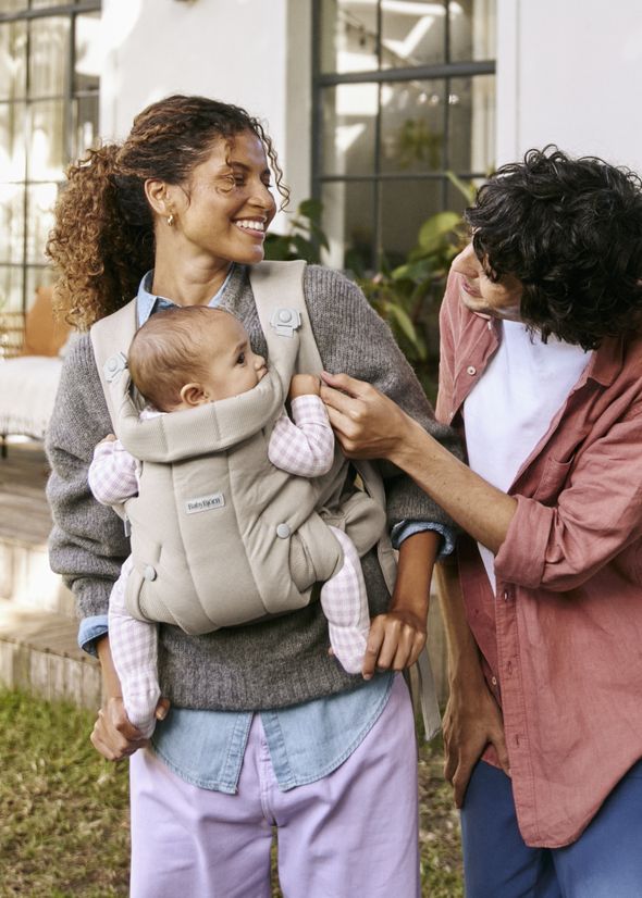 A smiling couple outdoors, with one wearing a baby carrier holding a baby, and the other adjusting the carrier strap.