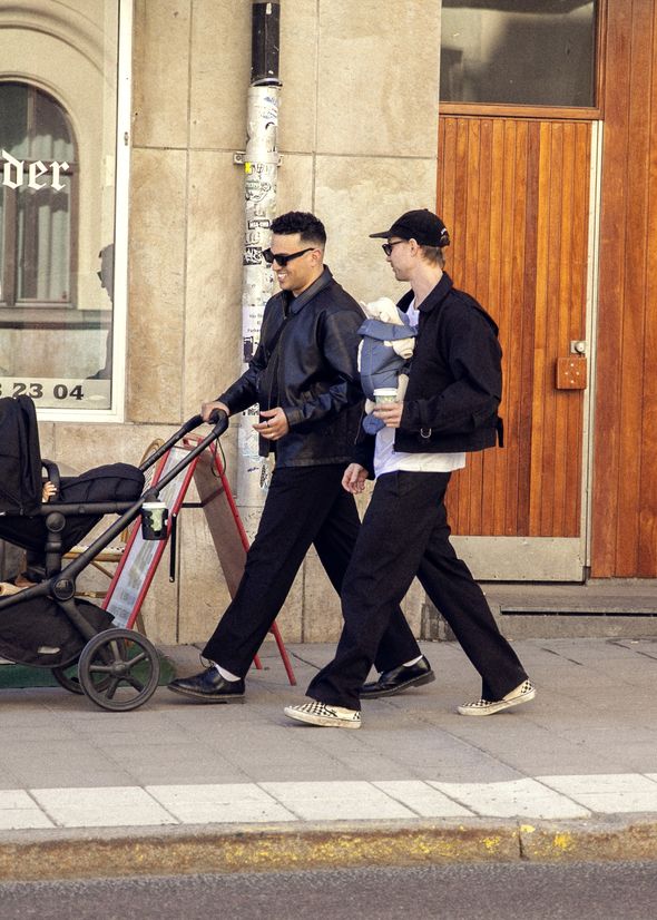 Two men walking on a sidewalk, one pushing a stroller, the other carrying a baby in a carrier, both wearing sunglasses and casual clothing.
