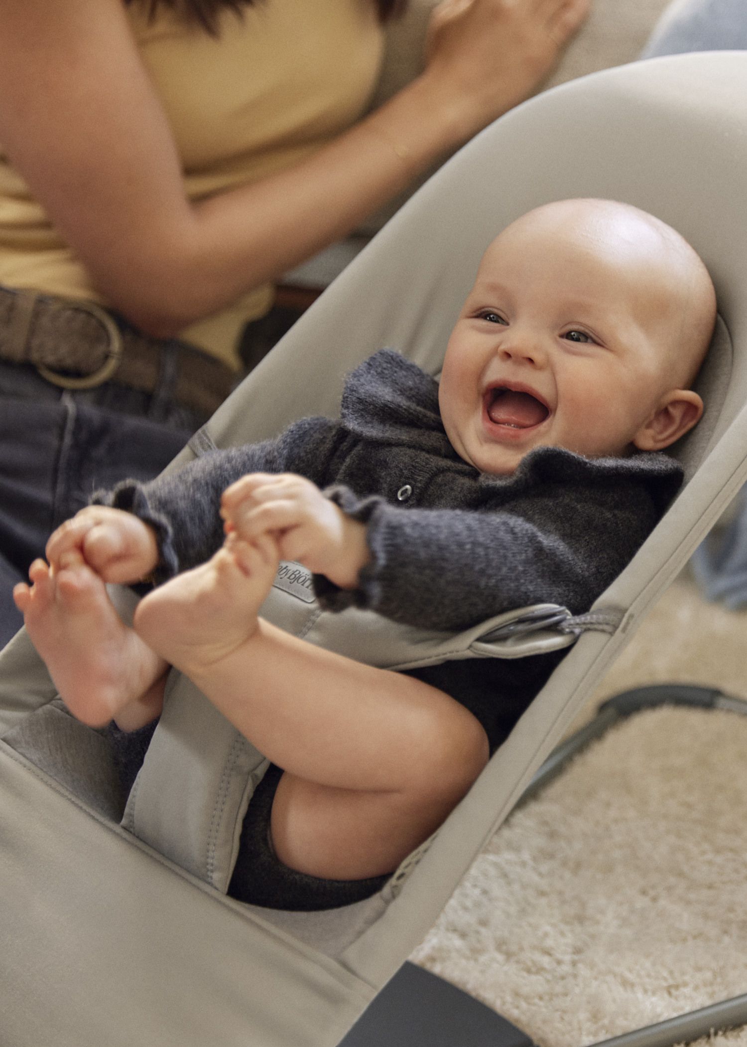 A baby in a gray outfit laughs joyfully while sitting in a baby bouncer. An adult is partially visible in the background.