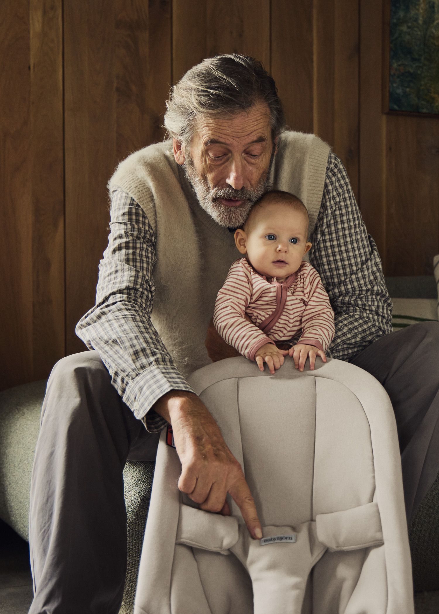An elderly man with a beard holds a baby in his lap, pointing to a label on a baby seat. They are indoors with wooden paneling in the background.