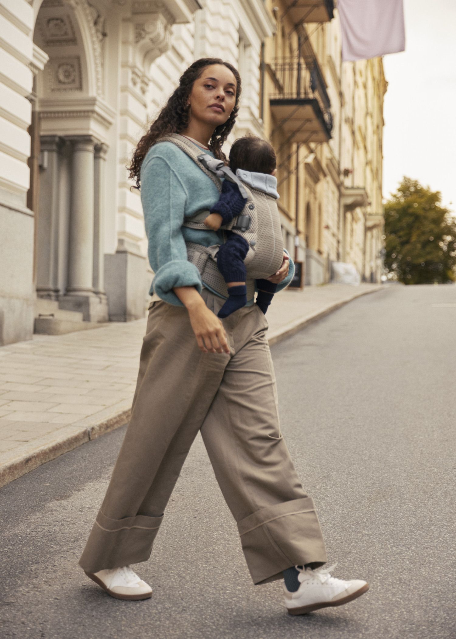 Woman walking on a city street, wearing a blue sweater and beige pants, carrying a baby in a front carrier.