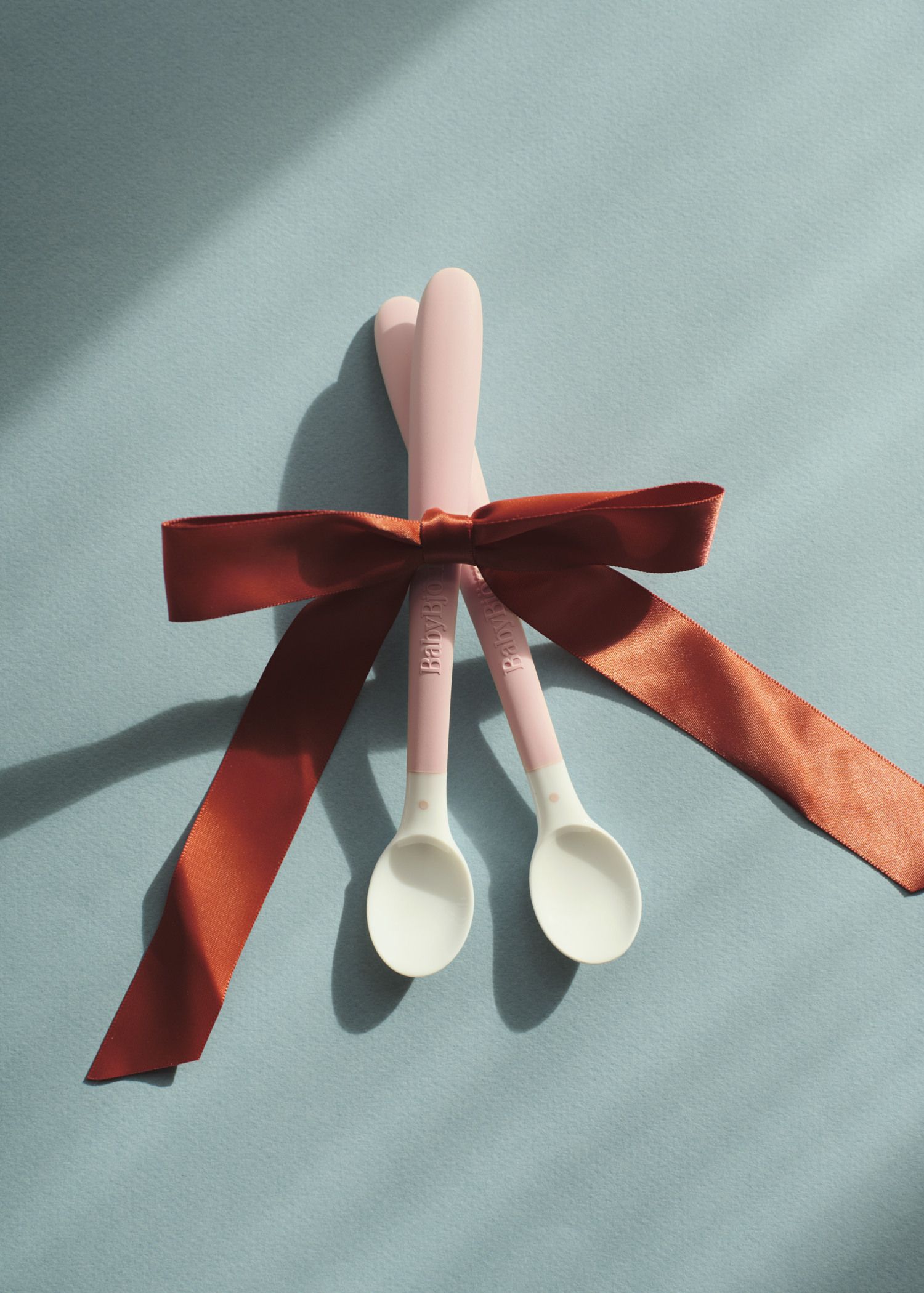 Two pink baby spoons tied together with a red ribbon on a light blue background, casting soft shadows.