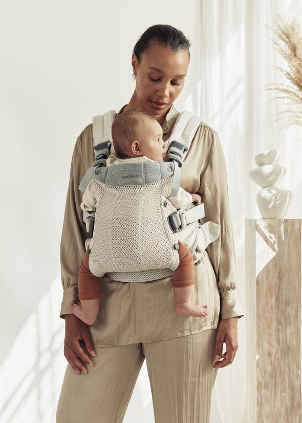 A woman in a beige outfit carries a baby in a mesh baby carrier. The room is softly lit with neutral decor and pampas grass in the background.