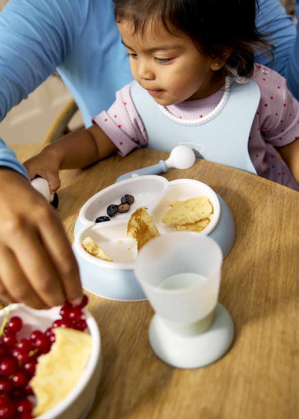 Toddler in a bib at a wooden table with a divided plate of pancakes and blueberries, adult hand reaching for a bowl of red berries and a silicone cup nearby.