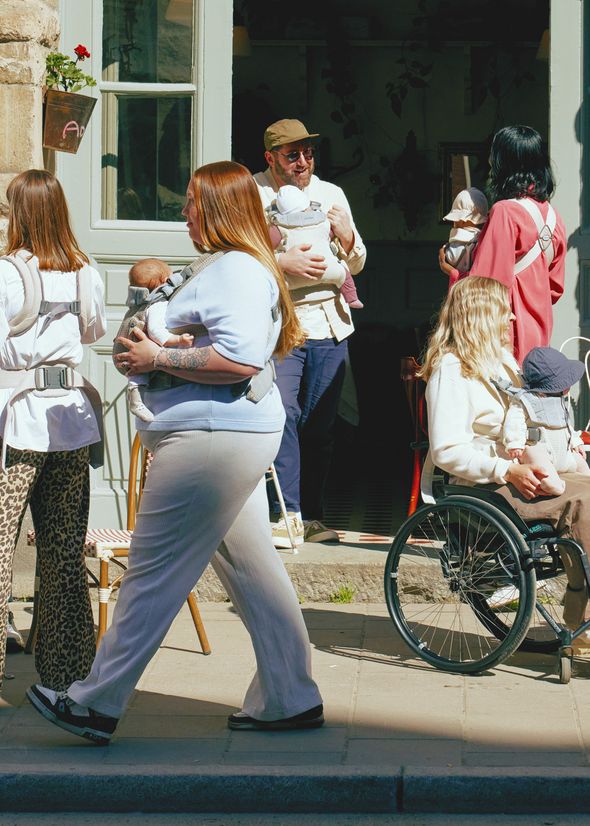 A group of people, some with babies in carriers, gather outside a building. One person is in a wheelchair. Sunlight casts shadows on the scene.