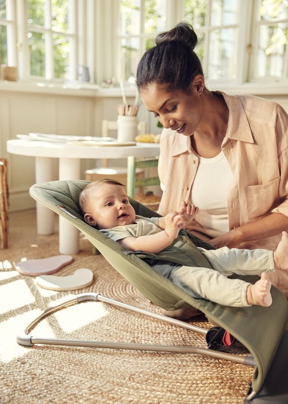 Mother smiling at her baby lying in a green bouncer. Bright room with natural light and wooden furniture in the background.