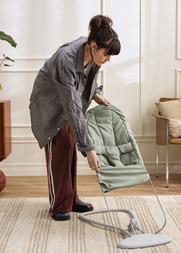 A person adjusts a green baby bouncer in a living room with wooden floors, striped pants, and a casual jacket.