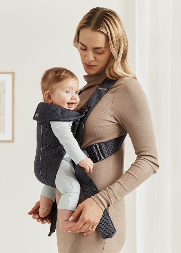A woman in a beige dress holds a smiling baby in a dark blue baby carrier, standing in a softly lit room.