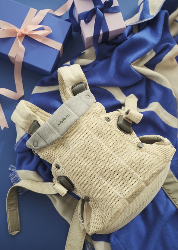A beige baby carrier on a blue and white striped blanket, surrounded by blue and pink gift boxes with ribbons.