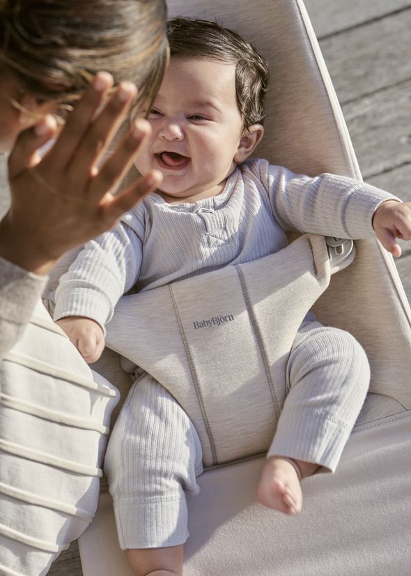 A baby wearing a light-colored outfit is sitting in a baby carrier, smiling and reaching towards a person who is playfully covering their face.
