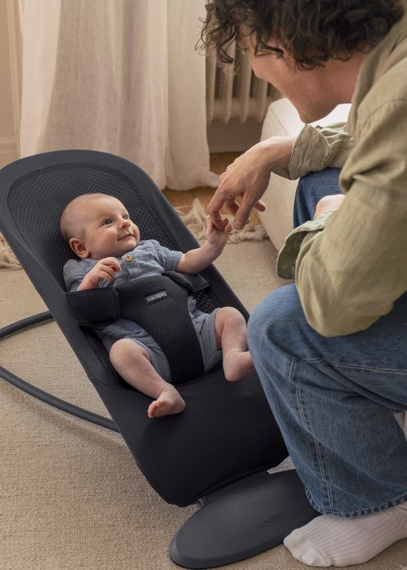 Parent kneeling beside a baby in a bouncer seat, holding the baby's hand, with the baby smiling back.