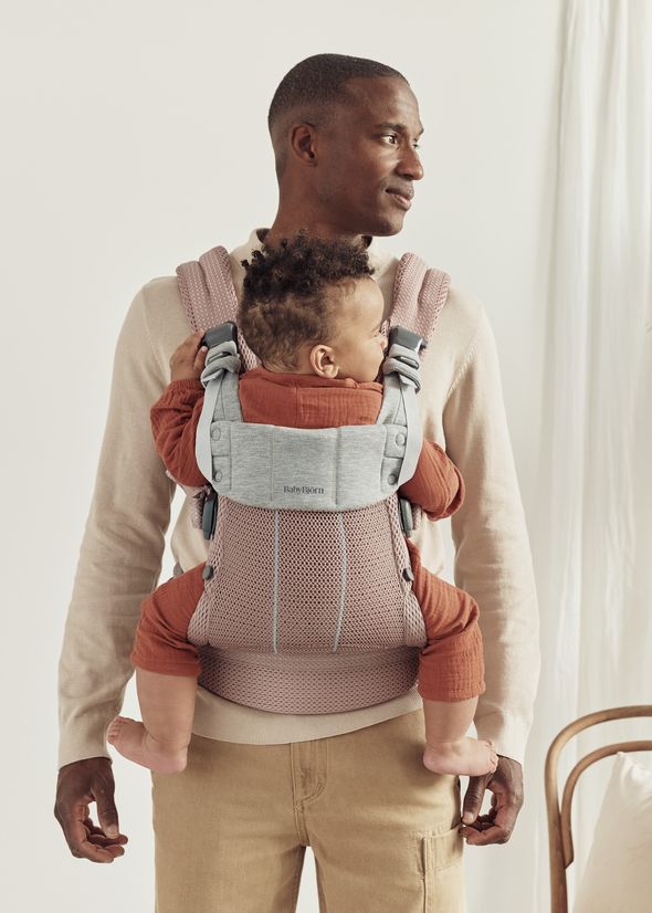 A man wearing a beige sweater carries a baby facing forward in a pink and gray baby carrier. The baby wears a rust-colored outfit.