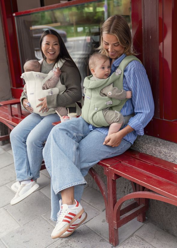 Two people sitting on a red bench, each wearing a baby carrier with a baby. They are smiling and dressed casually.