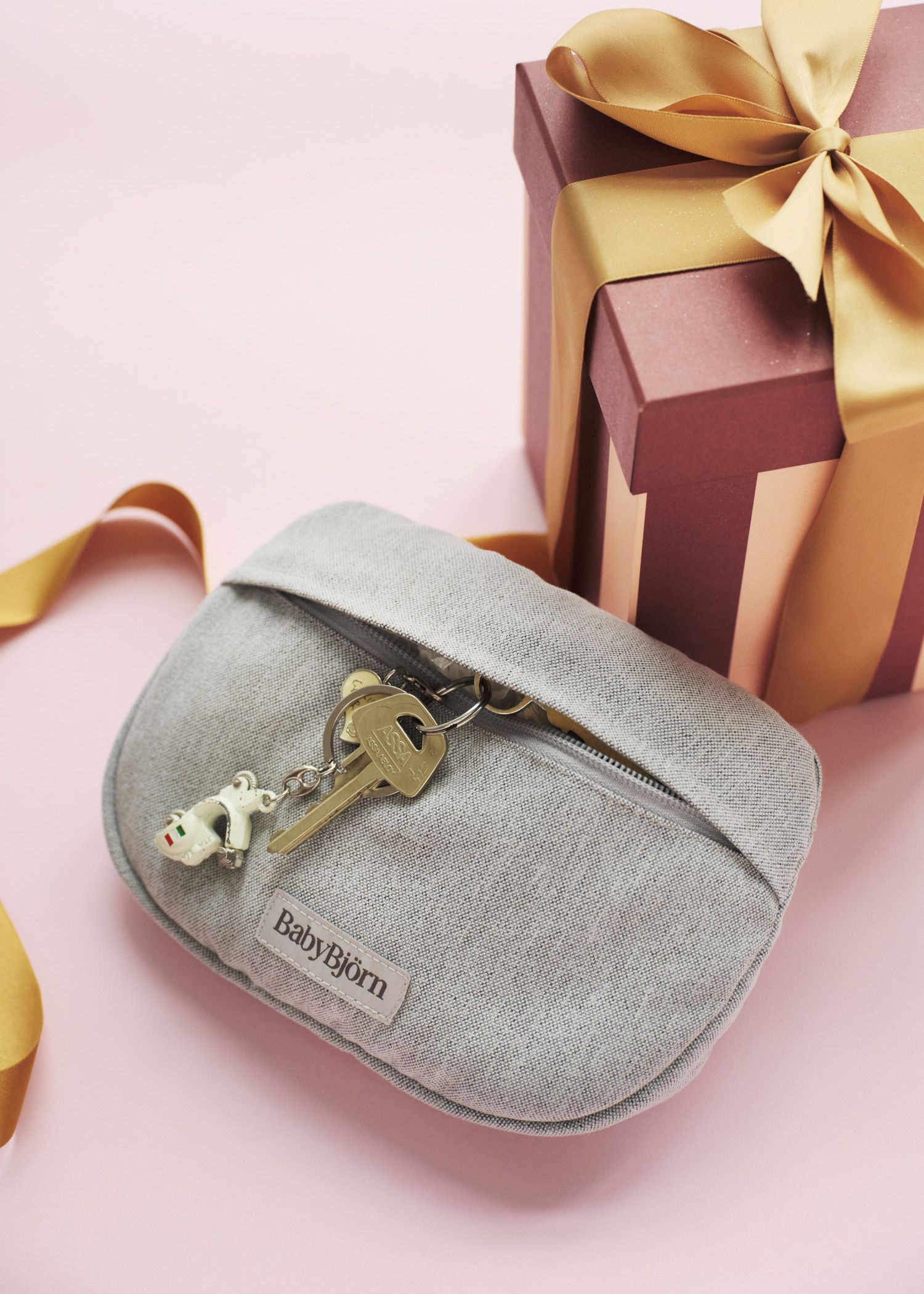 Gray BabyBjörn pouch with a keychain and keys on top, next to a pink and gold gift box with a ribbon on a pink background.