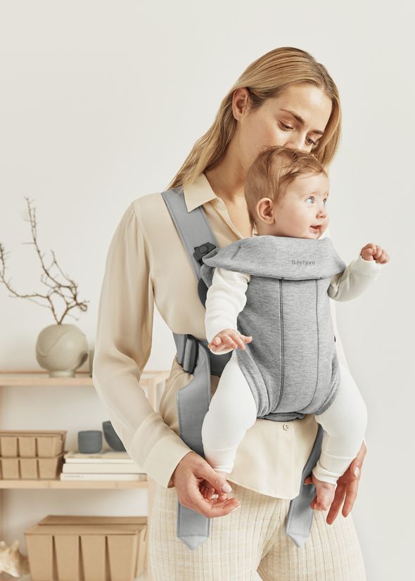 A woman wearing a beige blouse holds a baby in a gray baby carrier, standing in a cozy, minimalist room with shelves in the background.