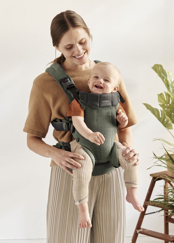 A smiling woman in a brown top carries a happy baby in a green mesh baby carrier. The setting is bright with a plant in the background.