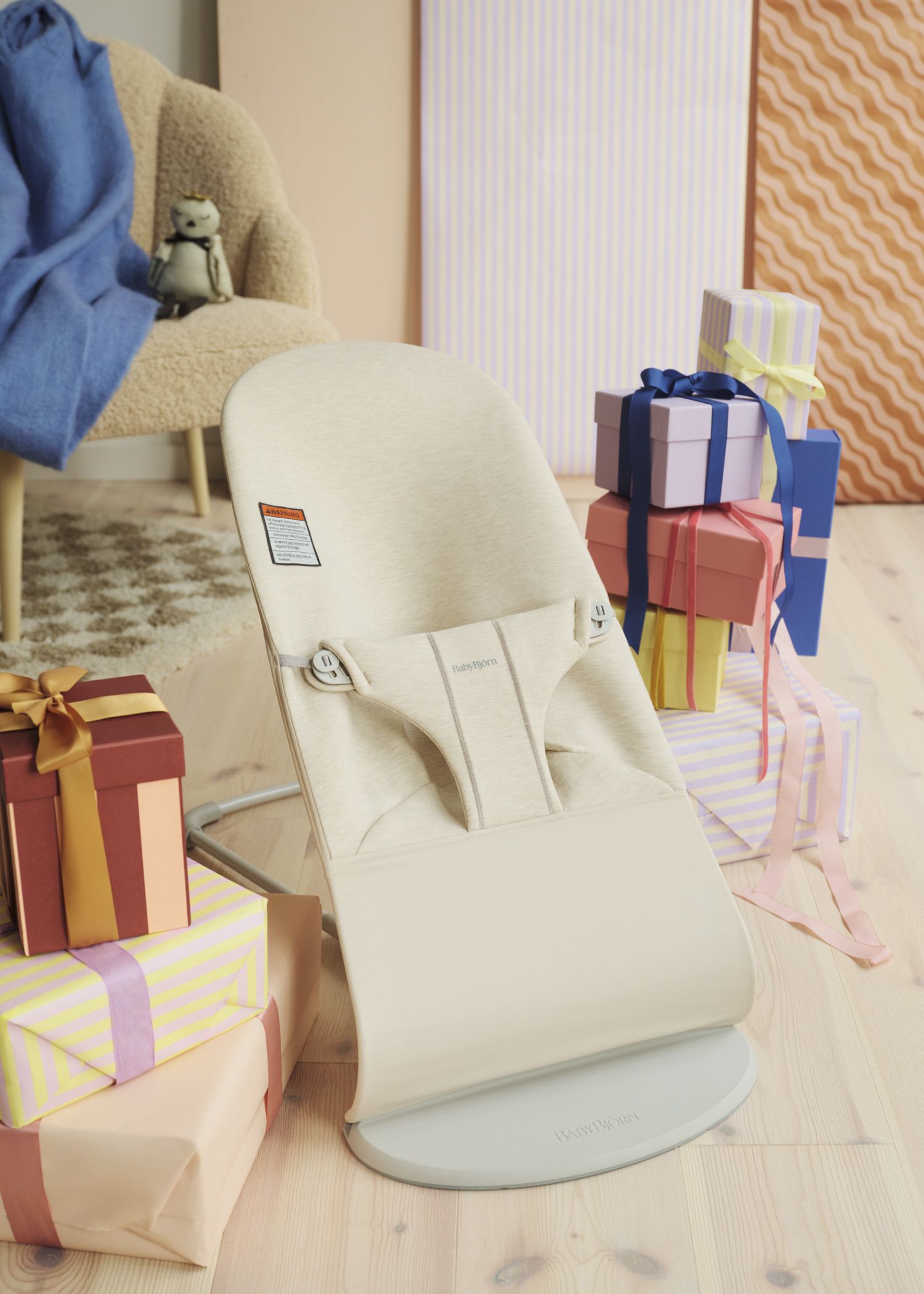 Baby bouncer surrounded by colorful, wrapped gifts on a wooden floor, with a cozy chair and a blue blanket in the background.