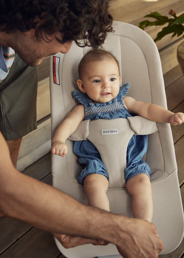 A smiling baby in a blue outfit sits in a gray baby bouncer, while an adult gently holds their foot, with a plant in the background.