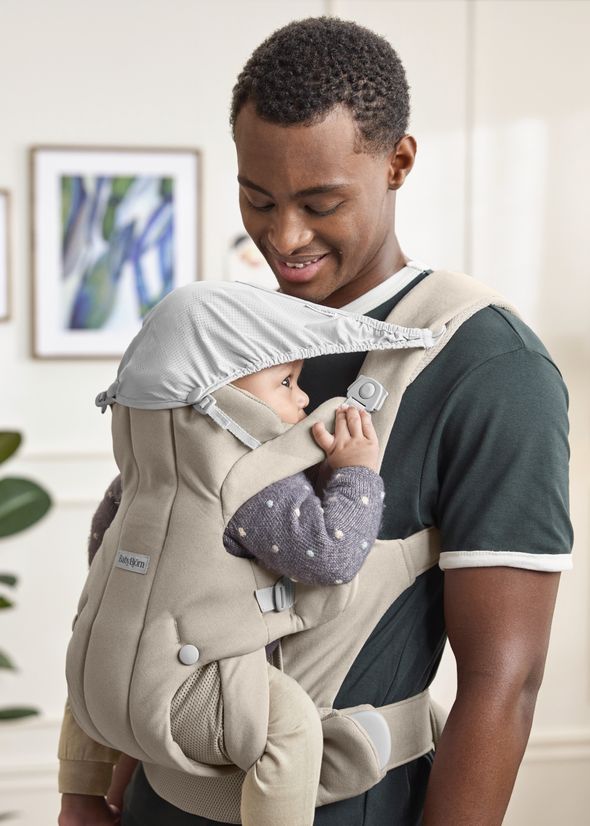 A man smiling at a baby in a beige baby carrier with a sunshade, standing indoors with artwork blurred in the background.