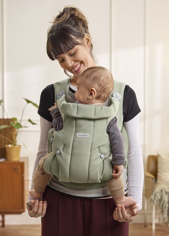 A smiling woman holds a baby in a soft green carrier in a cozy room, surrounded by plants and furniture.