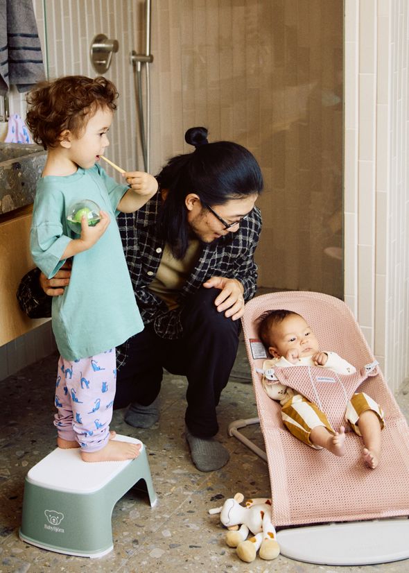 A person crouches beside a toddler brushing teeth on a stool, while a baby sits in a pink bouncer nearby in a bathroom setting.