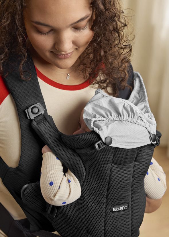 Woman smiling at a baby nestled in a black carrier. The baby wears a light gray hood and a cream outfit with blue dots.