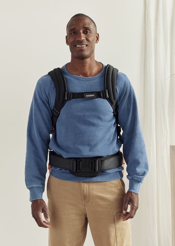 Man in blue sweatshirt wearing a black baby carrier over his shoulders and waist, standing in a brightly lit room.