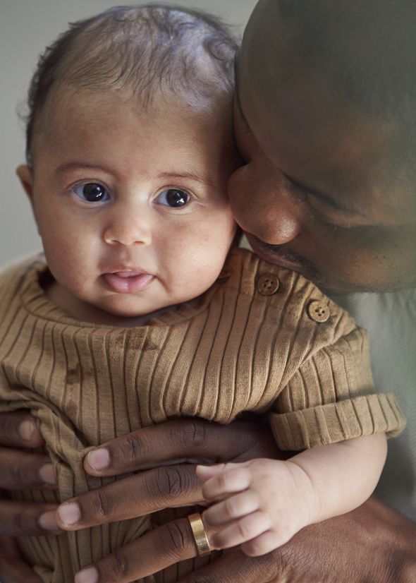 A man gently kisses a baby's cheek while holding the baby close, who is wearing a brown ribbed outfit and looking at the camera.