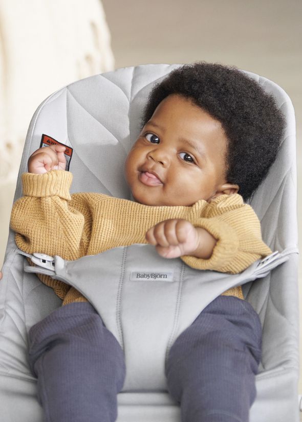 A baby with curly hair sits in a gray baby chair, wearing a mustard sweater and dark pants, looking up with a curious expression.