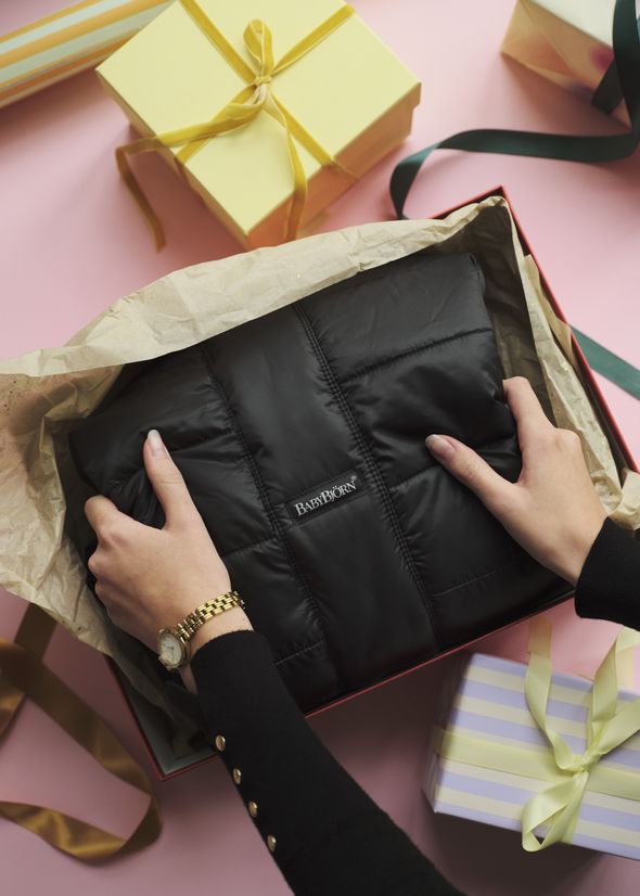 Hands holding a black padded item in a gift box, surrounded by wrapped presents on a pink surface.
