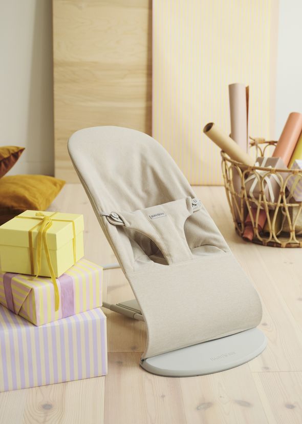 A beige baby bouncer on a light wooden floor, next to wrapped gifts in pastel colors and a basket with rolled papers.