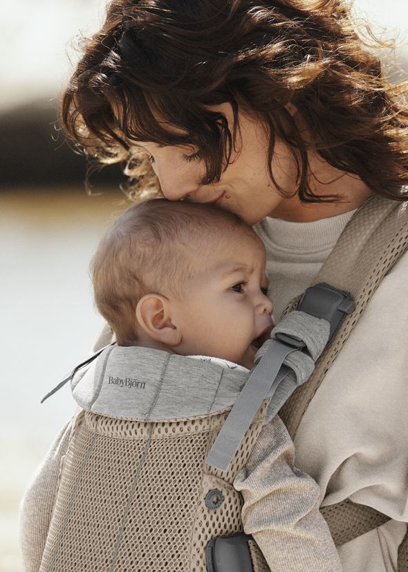 Mother gently kisses her baby's head while carrying them in a gray BabyBjörn carrier outdoors.