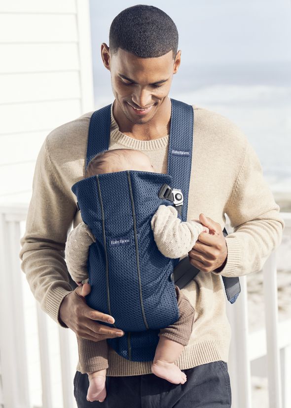 A person smiles while carrying a baby in a blue baby carrier. They are standing outside with a blurred background of a beach and building.