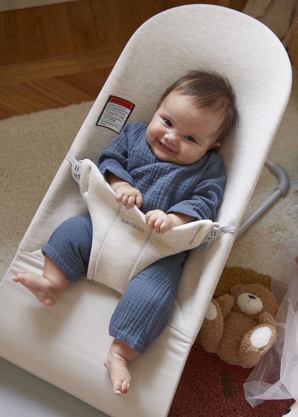 A smiling baby in a blue outfit sits in a cream-colored baby bouncer. A plush teddy bear is on the floor nearby.