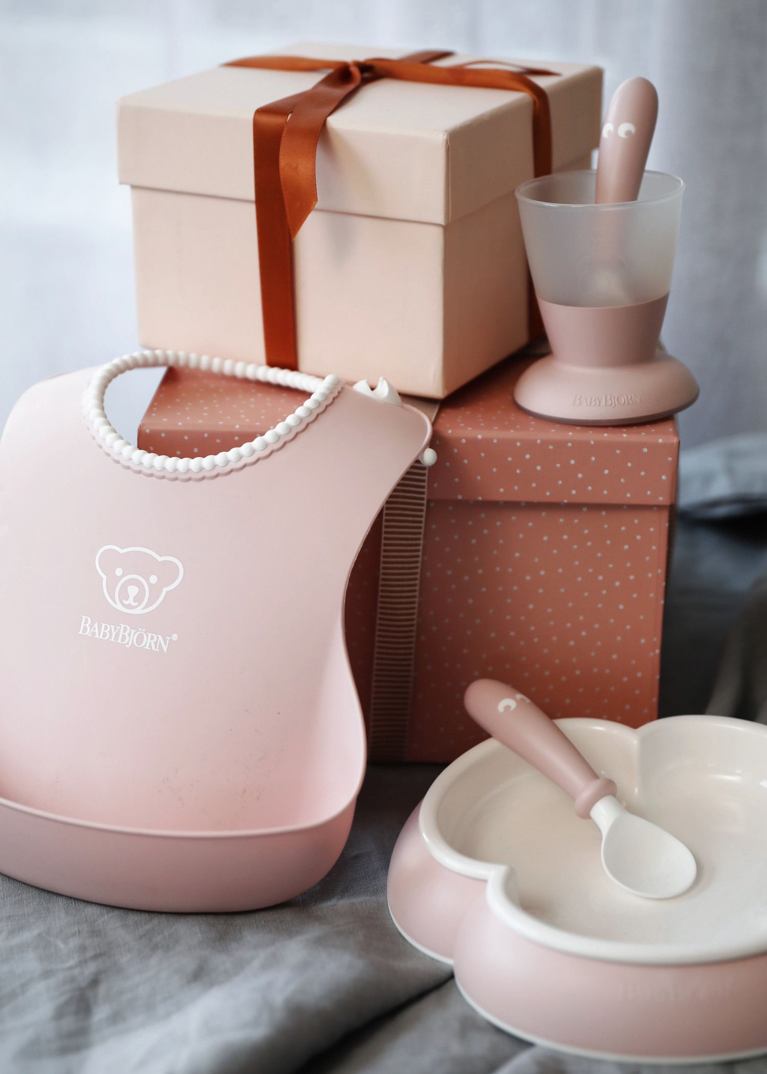 Pink baby dining set with bib, bowl, spoon, and cup, placed on stacked gift boxes with a brown ribbon.