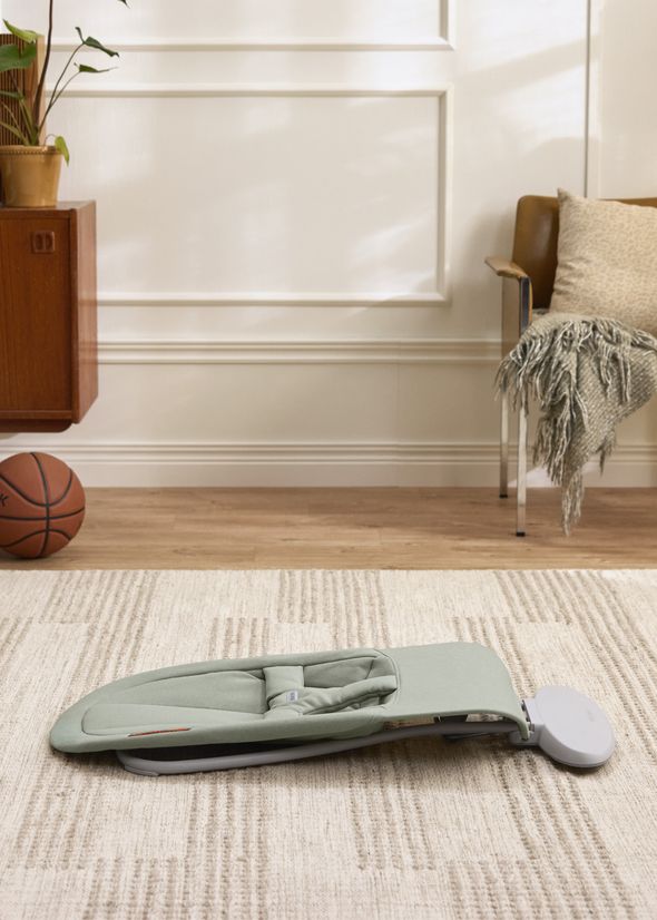 A pale green baby rocker is placed on a beige rug in a cozy room with a basketball, wooden cabinet, and an armchair with a throw blanket.