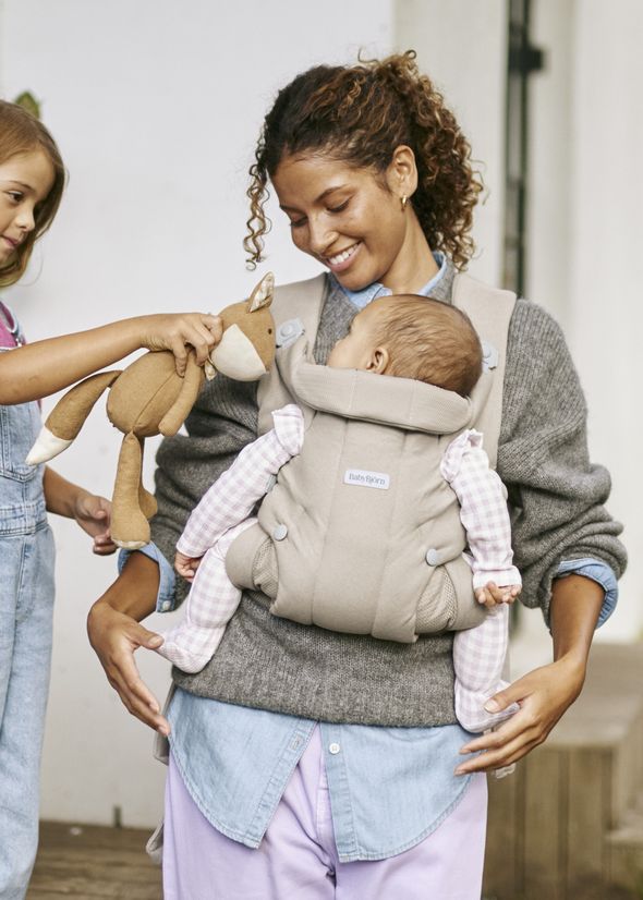 A smiling woman carries a baby in a beige carrier. A child offers a plush toy to the baby, who is looking at it.
