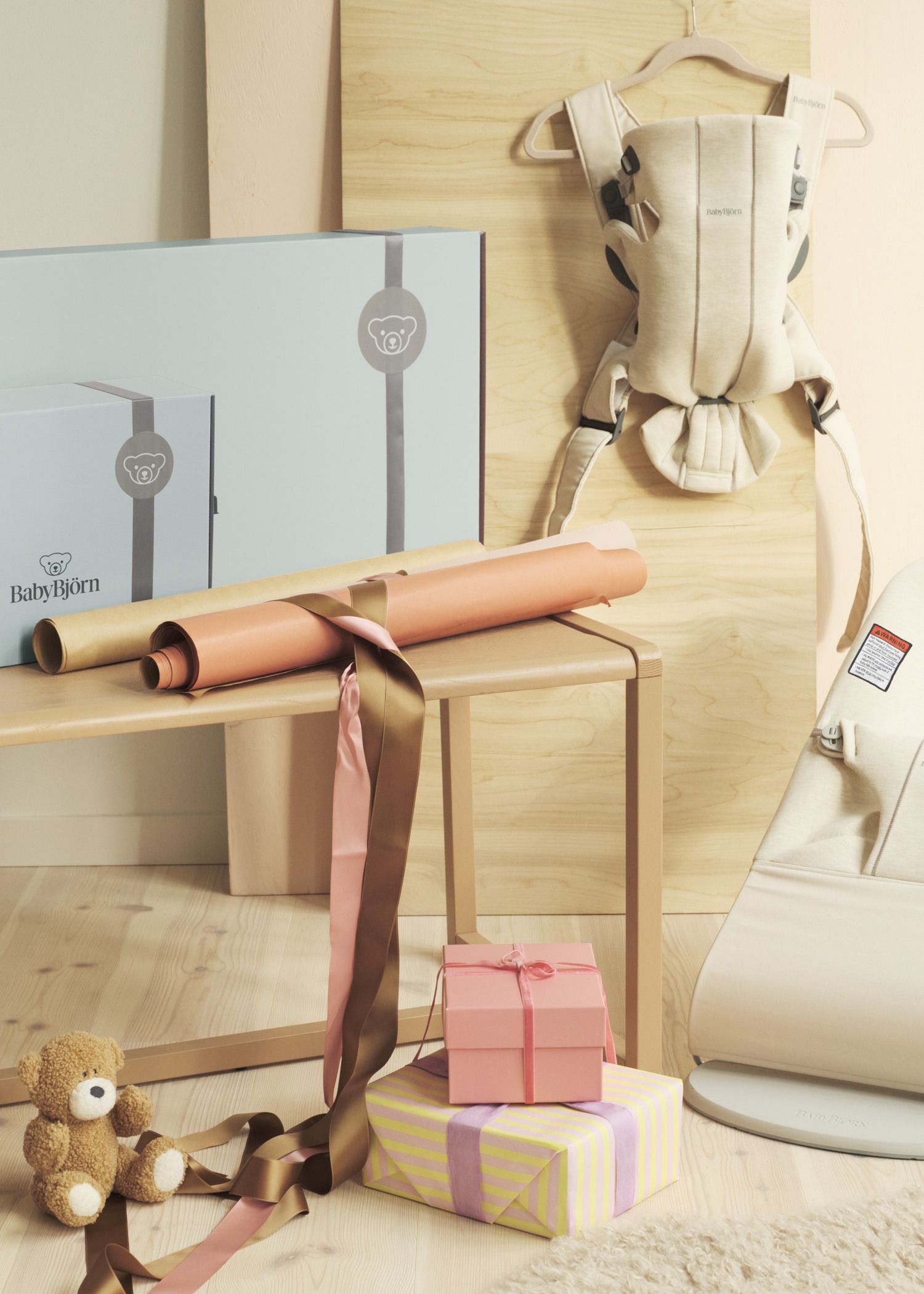 Gift-wrapped boxes, a teddy bear, and a baby carrier on a wooden table with decorative paper and ribbons.