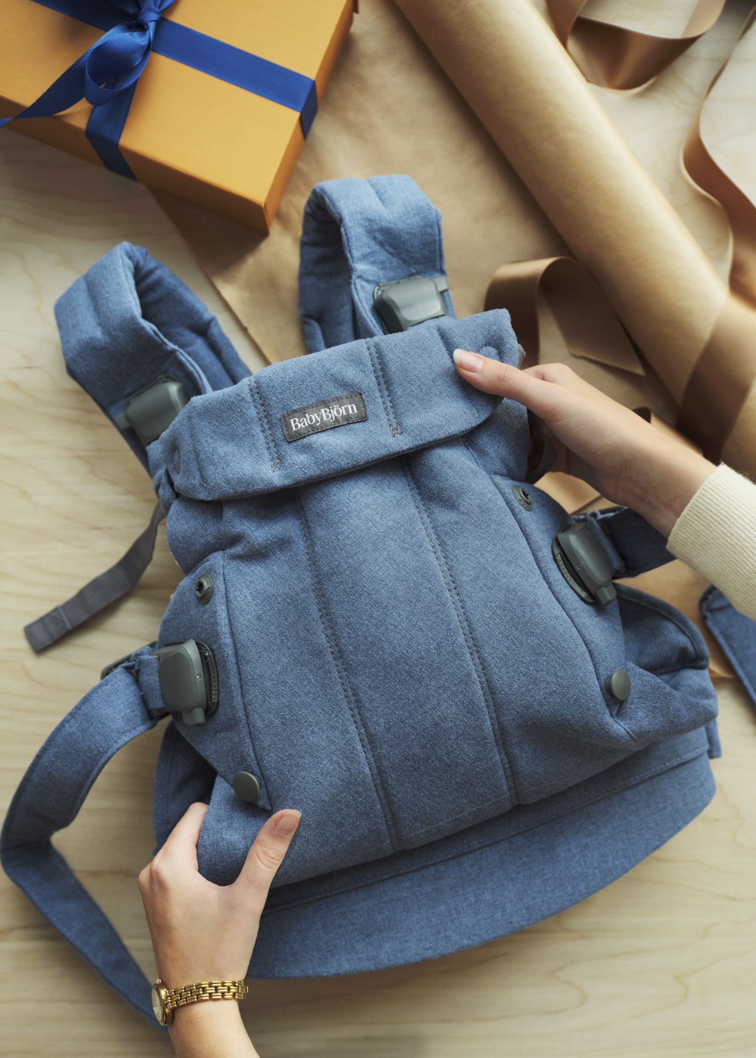 Hands holding a blue baby carrier on a wooden surface, with a wrapped gift and brown paper in the background.
