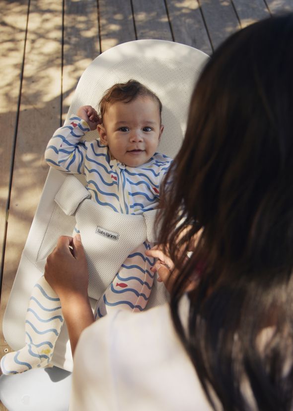 Baby in a patterned onesie lying in a bouncer chair, smiling up at an adult with hands gently holding the bouncer's straps.
