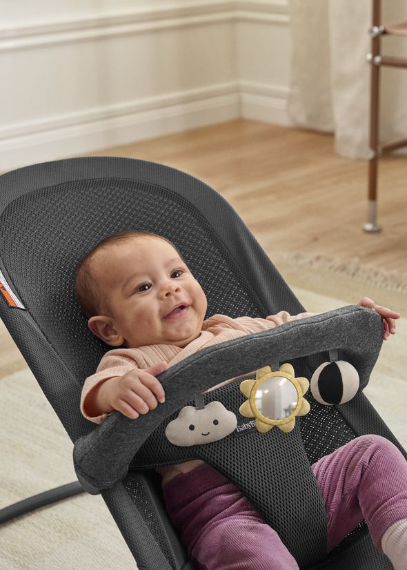 A smiling baby in a pink outfit sits in a dark gray baby bouncer with plush toys attached to the front bar in a bright room.