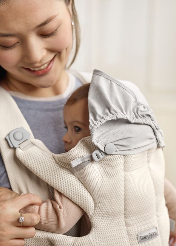 A smiling woman holds a baby in a beige BabyBjörn carrier. The baby, wearing a hood, is nestled comfortably against her chest.
