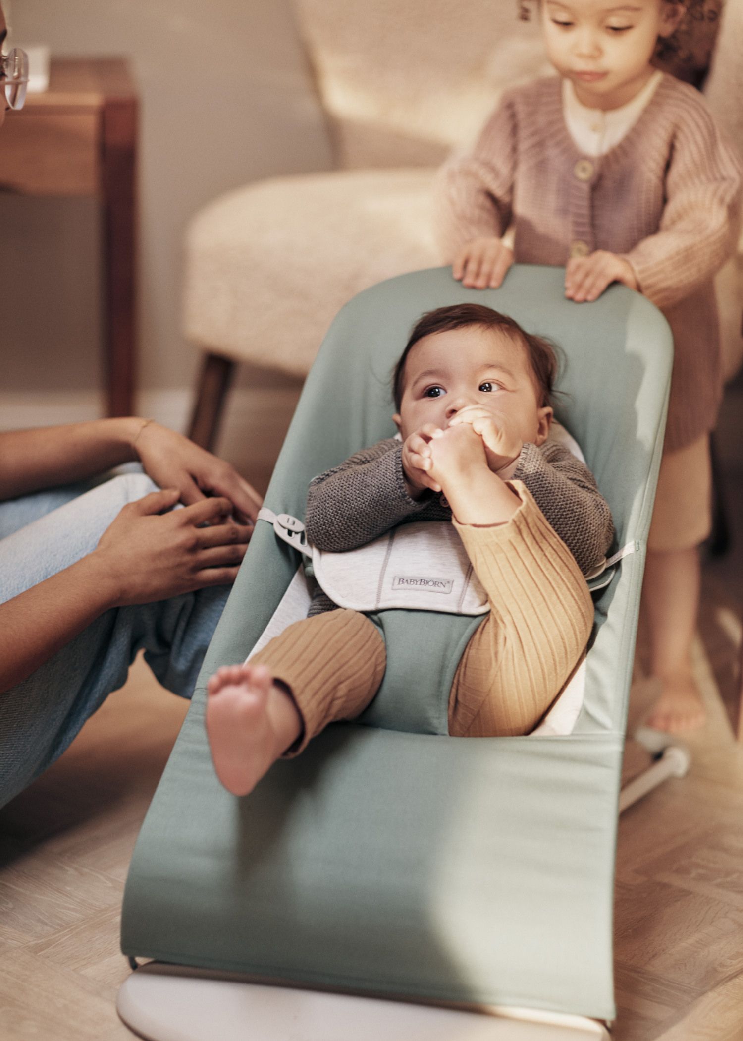 Baby in a green bouncer chair, holding a bottle, with a toddler standing nearby and an adult seated close, in a cozy room setting.