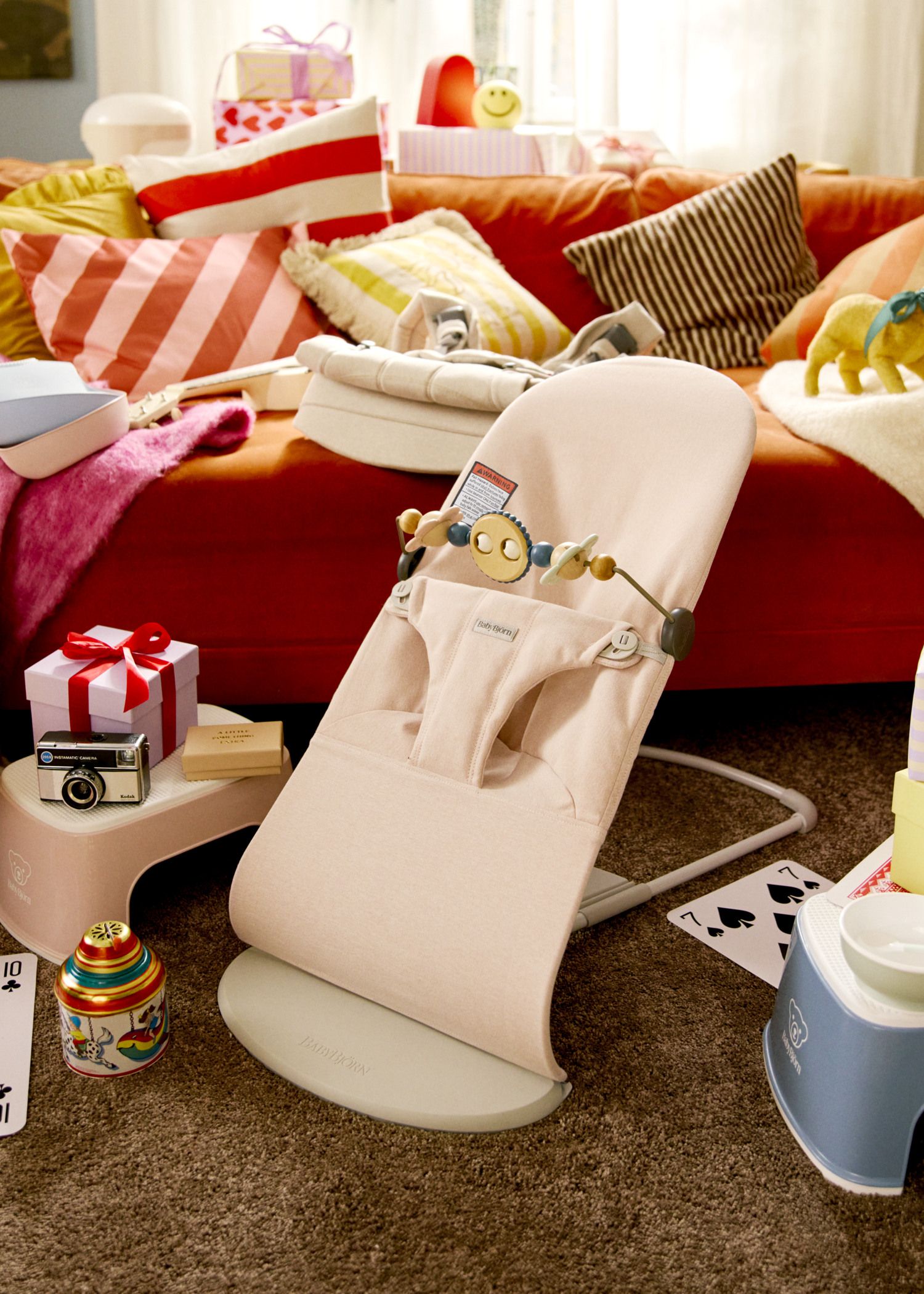 A cozy living room with a pink baby bouncer, toys, a camera, and gifts on a brown carpet. A red sofa with colorful pillows is in the background.