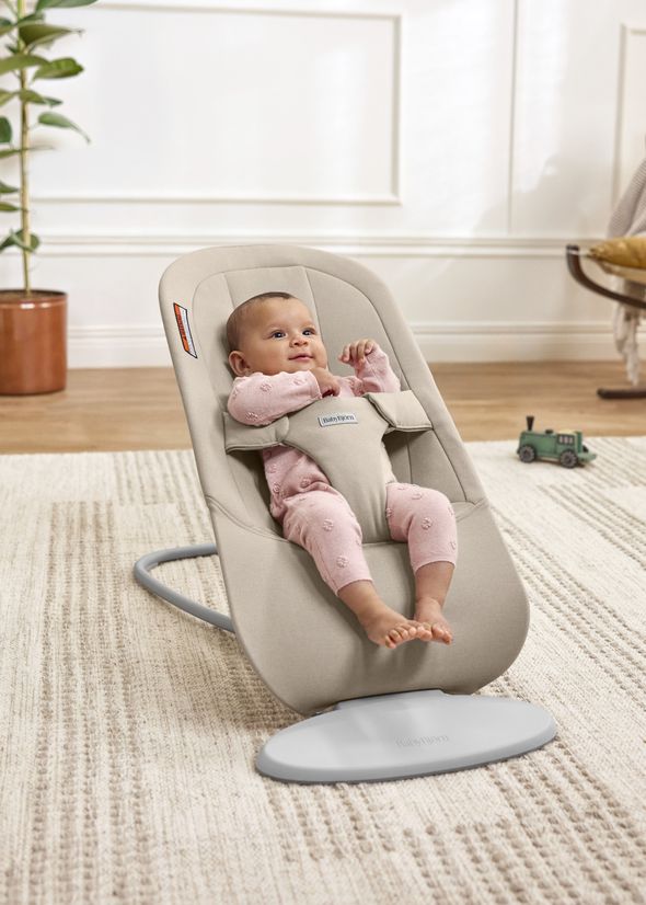 Baby in pink outfit smiling in a beige baby bouncer on a rug, green toy train in background, plant to the side.