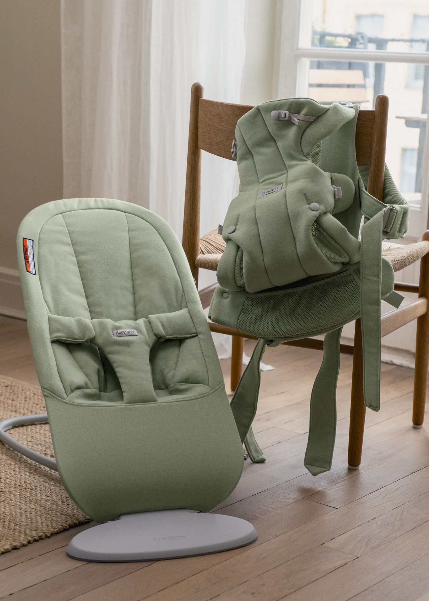 A green baby bouncer and ergonomic baby carrier are displayed on a wooden floor near a chair in a softly lit room.