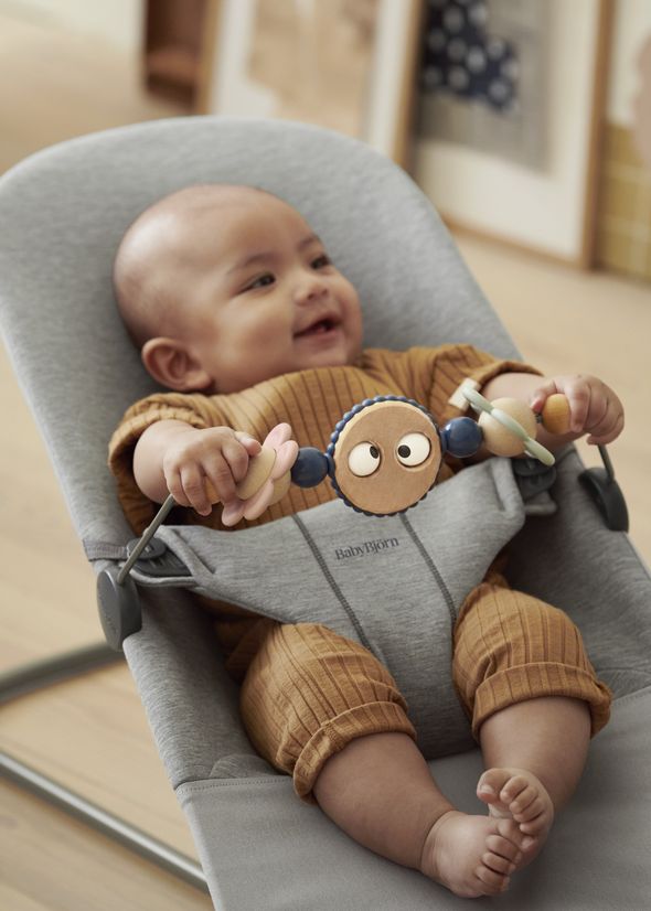 A smiling baby in a brown outfit sits in a gray bouncer holding a toy with colorful beads and faces.