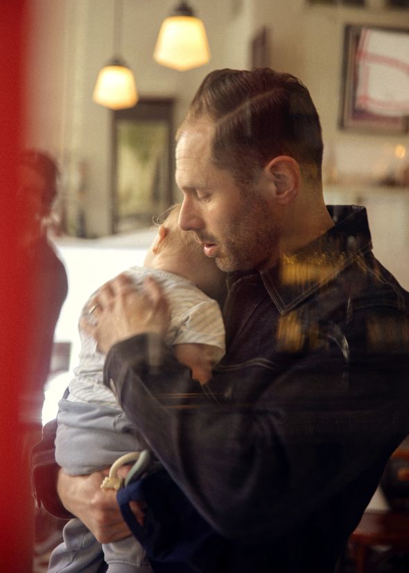 A man gently holds a sleeping baby against his shoulder in a cozy, softly lit room with hanging lights and blurred reflections in the window.